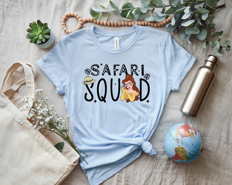 K&ouml;nnte beinhalten: Hellblaues T-Shirt mit dem Schriftzug "SAFARI SQUAD" in schwarzer Schrift und einer Cartoon-Illustration einer Frau mit Brille und gelbem Kleid. Ein Safarihut ist ebenfalls abgebildet.