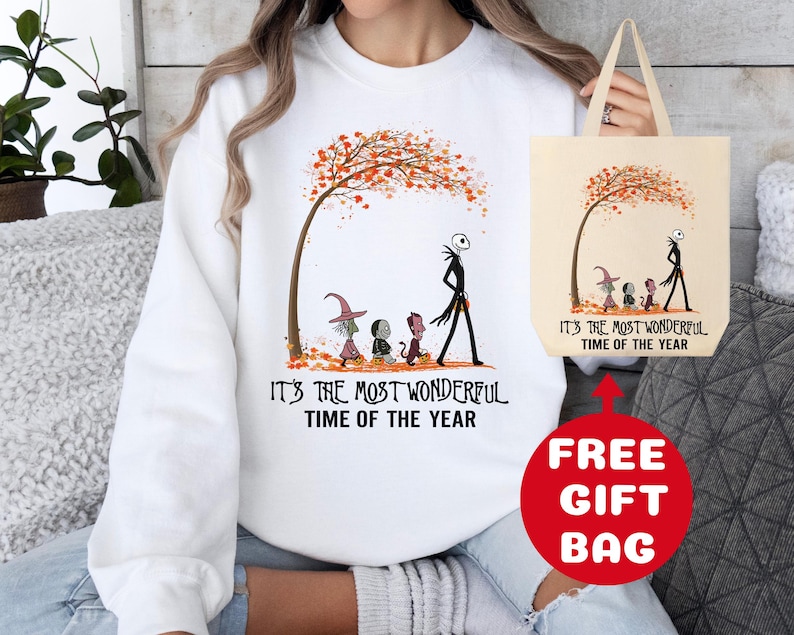 Op de afbeelding: Witte trui met een afbeelding van Jack Skellington en andere personages uit The Nightmare Before Christmas die door een herfstsc&egrave;ne lopen. De tekst "It's the most wonderful time of the year" staat op de trui gedrukt. Een gratis tote bag met dezelfde afbeelding en tekst is inbegrepen.