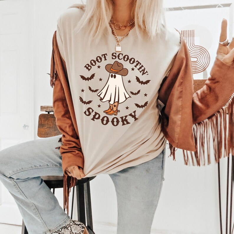 May include: A beige t-shirt with a graphic of a ghost wearing a cowboy hat and boots. The text "Boot Scootin' Spooky" is printed on the shirt.