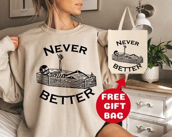 May include: A beige sweatshirt with the text "NEVER BETTER" and a graphic of a skeleton lying in a coffin. The sweatshirt comes with a free beige tote bag with the same graphic and text.