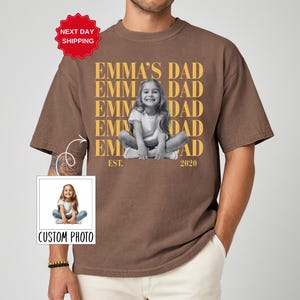 Puede incluir: Camiseta marr&oacute;n con el texto "EMMA'S DAD" repetido verticalmente en dorado, con una foto en blanco y negro de un ni&ntilde;o. La camiseta tambi&eacute;n tiene el texto "EST. 2020" y una etiqueta "CUSTOM PHOTO".