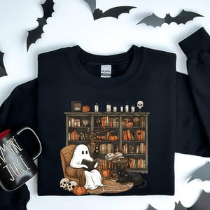May include: Black sweatshirt with a graphic of a ghost reading a book in a library with a black cat. The text "Vampire Juice" is on a black mug.