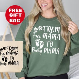 May include: A green sweatshirt with white text that reads "From Fur Mama to Baby Mama" and paw prints. The sweatshirt is being modeled by a person. A tote bag with the same text and paw prints is also shown in the image.
