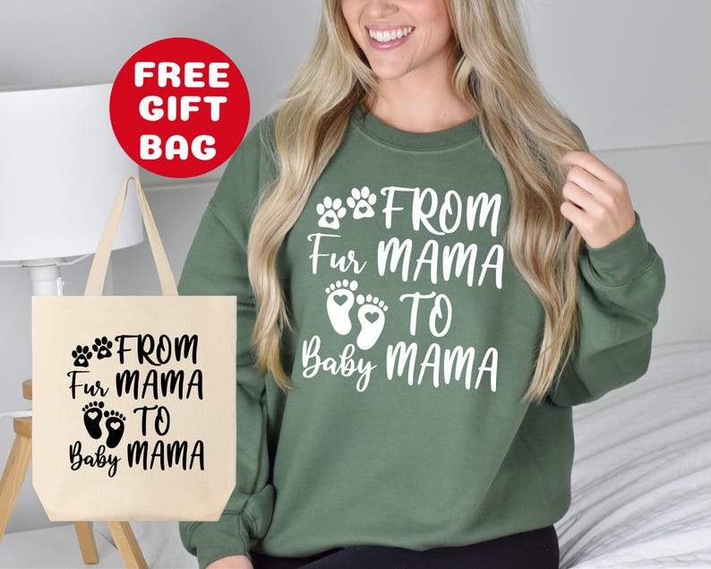 May include: A green sweatshirt with white text that reads "From Fur Mama to Baby Mama" and paw prints. The sweatshirt is being modeled by a person. A tote bag with the same text and paw prints is also shown in the image.