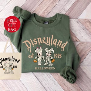 May include: Green sweatshirt with a Disneyland Halloween design. The design features Mickey Mouse and Minnie Mouse dressed as mummies holding pumpkins. The text reads "Disneyland est. 1928 Halloween".