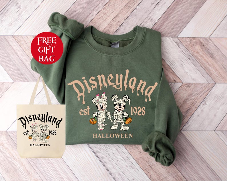 May include: Green sweatshirt with a Disneyland Halloween design. The design features Mickey Mouse and Minnie Mouse dressed as mummies holding pumpkins. The text reads "Disneyland est. 1928 Halloween".