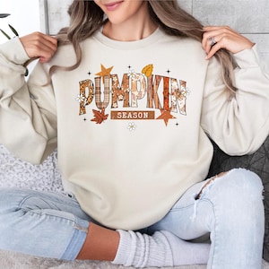 Może przedstawiać: Kremowa bluza z napisem "PUMPKIN SEASON" w jesiennym motywie. Tekst zawiera pomarańczowe, brązowe i beżowe wzory oraz kwiatowe akcenty. Bluza ma długie rękawy i okrągły dekolt.