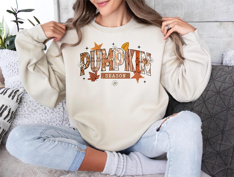 Może przedstawiać: Kremowa bluza z napisem "PUMPKIN SEASON" w jesiennym motywie. Tekst zawiera pomarańczowe, brązowe i beżowe wzory oraz kwiatowe akcenty. Bluza ma długie rękawy i okrągły dekolt.
