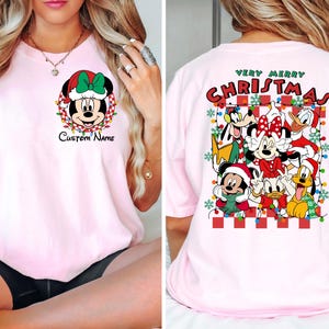 Op de afbeelding: Roze T-shirt met een kerstthema met Mickey Mouse en andere Disney-figuren. De voorkant van het shirt heeft een afbeelding van Mickey Mouse met een kerstmanmuts en de tekst "Custom Name". De achterkant van het shirt heeft de tekst "Very Merry Christmas" en een afbeelding van Mickey Mouse, Minnie Mouse, Donald Duck, Goofy, Pluto en Daisy Duck.