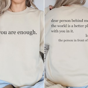 Puede incluir: Una sudadera beige con el texto "you are enough." impreso en la parte delantera y el texto "dear person behind me, the world is a better place with you in it. love, the person in front of you" impreso en la parte trasera.