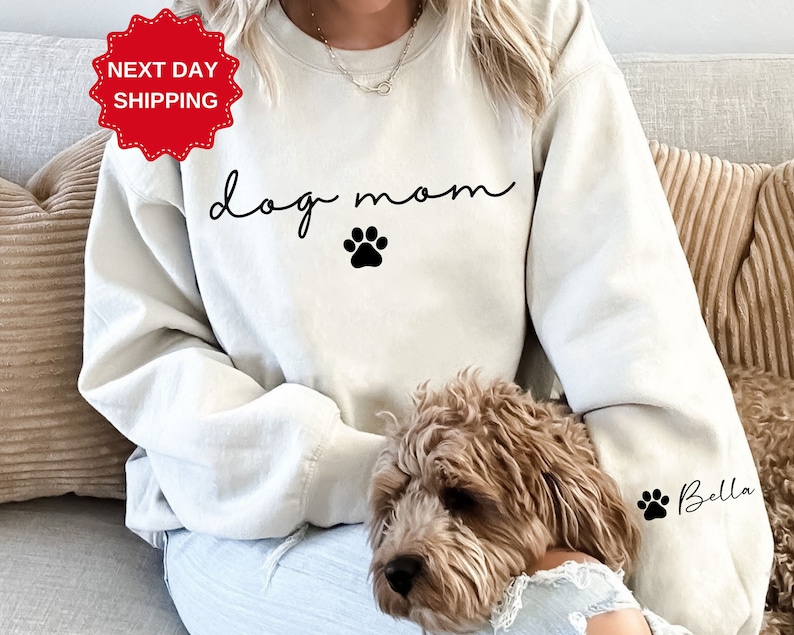 May include: A white sweatshirt with the text "dog mom" in black script and a black paw print. The sweatshirt is being worn by a person and a brown dog is sitting on their lap. The dog's name "Bella" is written in black on the sleeve of the sweatshirt.