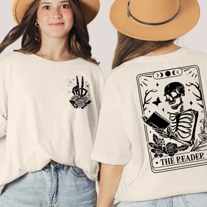 以下が含まれることがあります： 黒い骨格が本を読んでいる白いTシャツ。骨格の周りは花で飾られています。骨格の下には「THE READER」という文字があります。Tシャツの裏面には、黒い平和マークと花が描かれています。