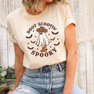 May include: A cream colored t-shirt with a graphic of a ghost wearing a cowboy hat and boots. The text "Boot Scootin' Spooky" is printed on the shirt.