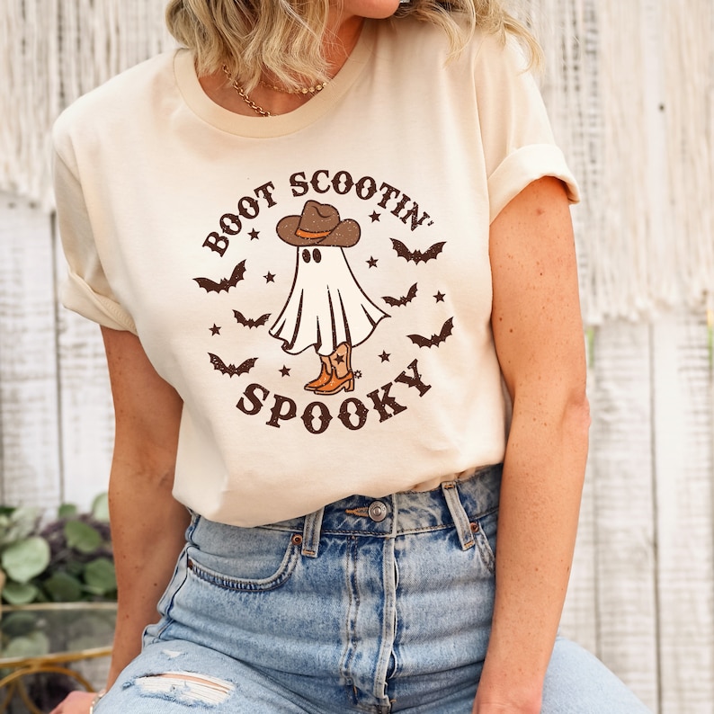May include: A cream colored t-shirt with a graphic of a ghost wearing a cowboy hat and boots. The text "Boot Scootin' Spooky" is printed on the shirt.