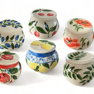May include: A collection of six small, round ceramic jars with lids. Each jar is hand-painted with a different fruit or botanical design, including cherries, lemons, pomegranates, olives, and poppies. The jars are white with colorful accents.
