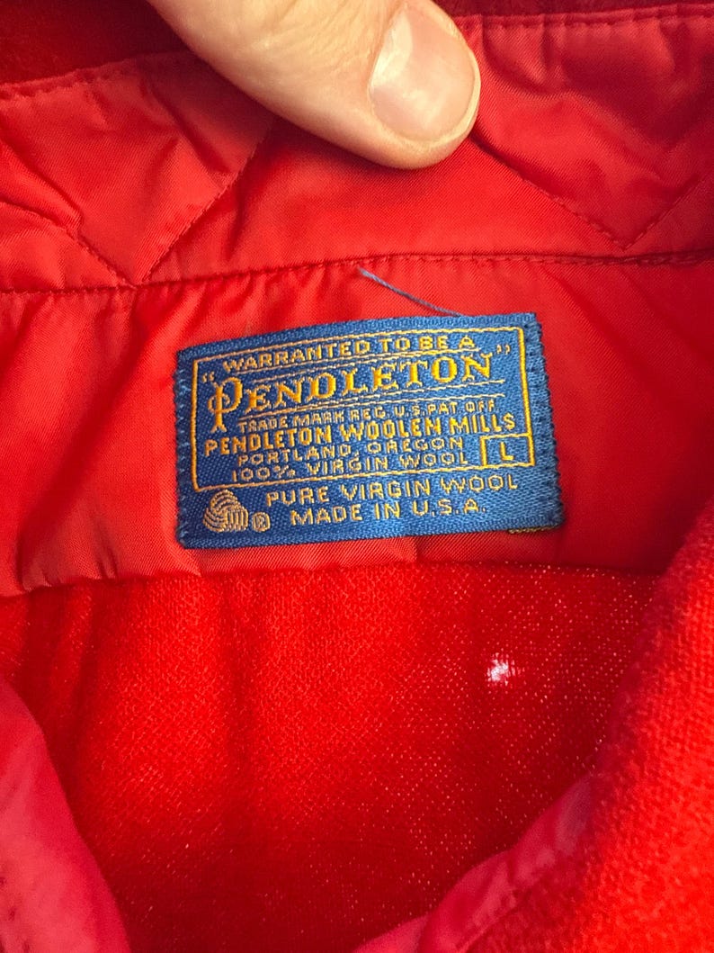 Puede incluir: Un interior de prenda acolchada de color rojo brillante con una etiqueta de tela azul rectangular con texto dorado que dice Warranted to be a Pendleton, Pendleton Woolen Mills, Portland, Oregon, 100% Virgin Wool, talla L y Made in U.S.A.