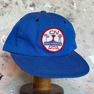 Puede incluir: Gorra de béisbol azul con visera curva. La gorra presenta un parche circular con el texto "CN ENGINEERING" en un diseño rojo y blanco. La gorra se exhibe sobre un soporte de madera.