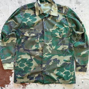 May include: A long-sleeved camouflage shirt in shades of green, brown, and black. The shirt features a button-down front, two chest pockets with button closures, and a classic collar. The fabric has a military-inspired pattern.