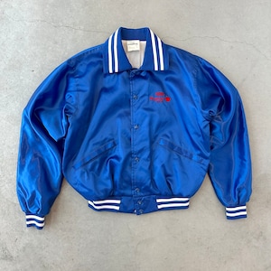 May include: A royal blue satin bomber jacket with a white collar and cuffs, accented with two white stripes. The jacket features a button-up front and a red logo on the chest. The jacket has a classic, vintage style.