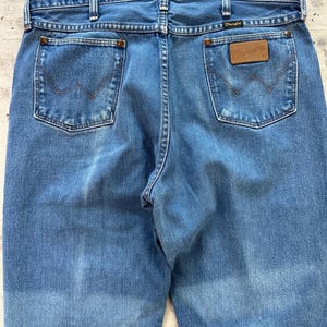 May include: Light blue denim jeans with classic five-pocket design. Features include copper rivets, a brown leather patch with the "Wrangler" logo, and a stitched back pocket design. The jeans have a faded appearance.