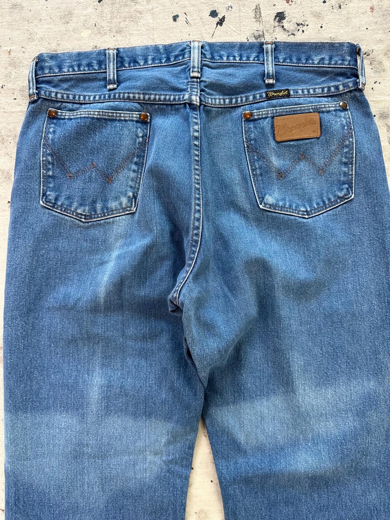 May include: Light blue denim jeans with classic five-pocket design. Features include copper rivets, a brown leather patch with the "Wrangler" logo, and a stitched back pocket design. The jeans have a faded appearance.