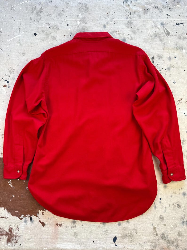 Puede incluir: Una camisa de manga larga con botones en un color rojo vibrante y s&oacute;lido. La camisa presenta un cuello cl&aacute;sico, un canes&uacute; en la espalda y pu&ntilde;os al final de cada manga, cada uno asegurado con un peque&ntilde;o bot&oacute;n. La tela parece tener una textura lisa.