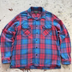 Puede incluir: Una camisa de cuadros de manga larga en tonos rojos, azules y morados. La camisa tiene cuello de campamento, dos bolsillos en el pecho y botones negros. El estampado de cuadros es un diseño clásico.