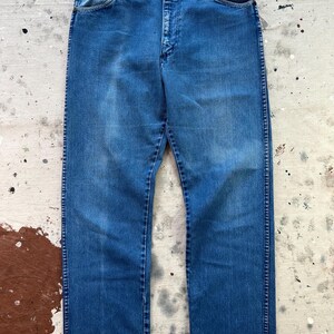 May include: A pair of vintage blue denim jeans. The jeans have a classic five-pocket design, a button and zipper closure, and a straight leg fit. The denim has a faded appearance, with lighter areas on the thighs and knees.