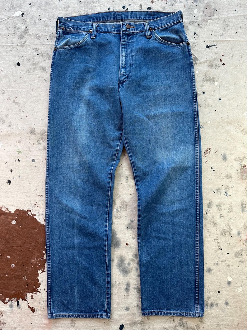 May include: A pair of vintage blue denim jeans. The jeans have a classic five-pocket design, a button and zipper closure, and a straight leg fit. The denim has a faded appearance, with lighter areas on the thighs and knees.