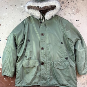 Op de afbeelding: Olijfgroene parka met een met bont gevoerde capuchon. De jas heeft een ritssluiting, knopen en twee voorzakken. De capuchon heeft een witte en bruine imitatiebont afwerking.