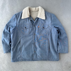 Op de afbeelding: Een blauwe denimjas met een sherpa voering en een knoopsluiting. De jas heeft twee klepzakken aan de voorkant en een rood label op de rechterzak.