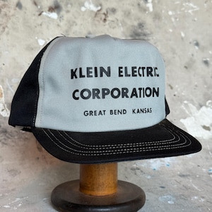 Könnte beinhalten: Schwarze und graue Trucker-Mütze mit dem Text "KLEIN ELECTRIC CORPORATION GREAT BEND KANSAS" auf der Vorderseite. Die Mütze hat eine graue Frontplatte und einen schwarzen Schirm und ein Mesh-Rückenteil. Die Mütze wird auf einem Holzständer ausgestellt.