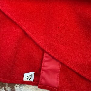 Puede incluir: Una prenda de tela roja vibrante con una textura suave y densa y un dobladillo cosido visible. Una peque&ntilde;a etiqueta rectangular blanca con texto negro parcial est&aacute; unida al borde inferior, junto a una tira vertical de acabado satinado rojo.