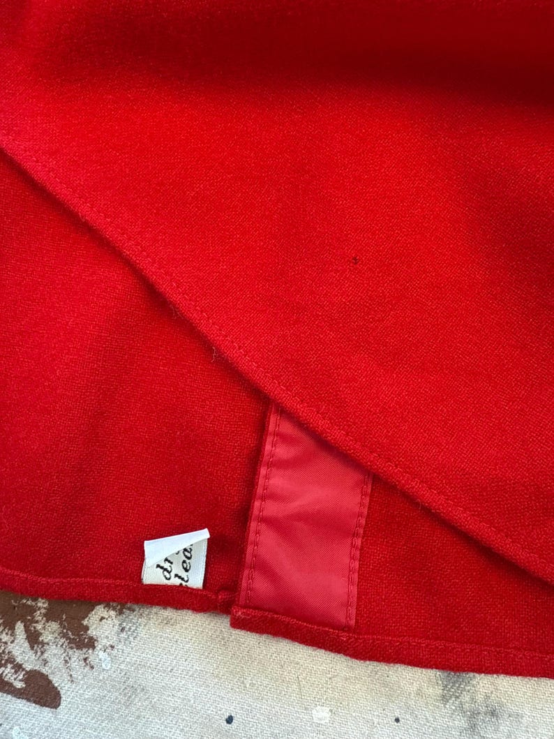 Puede incluir: Una prenda de tela roja vibrante con una textura suave y densa y un dobladillo cosido visible. Una peque&ntilde;a etiqueta rectangular blanca con texto negro parcial est&aacute; unida al borde inferior, junto a una tira vertical de acabado satinado rojo.