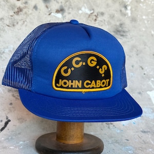 May include: A blue trucker hat with a mesh back and a curved brim. The hat features a black patch with the text "C.C.G.S JOHN CABOT" in yellow lettering. The hat is displayed on a wooden stand.