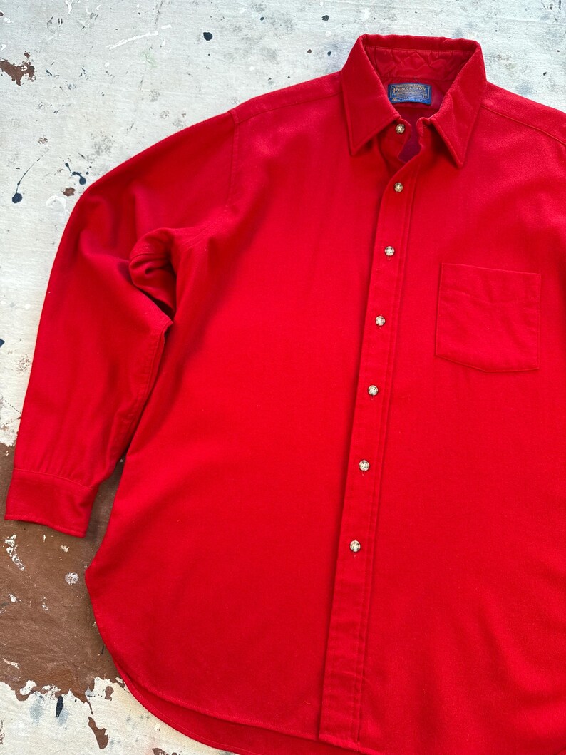 Puede incluir: Una camisa de manga larga en color rojo s&oacute;lido con cuello en punta, un bolsillo en el pecho y una serie de botones redondos de color claro en la parte frontal. La etiqueta interior del cuello muestra la marca Pendleton.