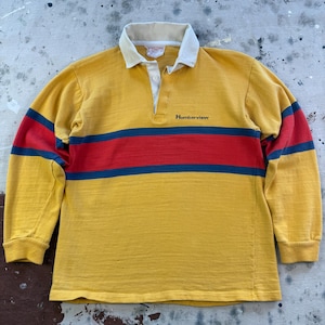 Puede incluir: Una camisa de rugby de manga larga en color amarillo mostaza con cuello color crema y tapeta de botones. La camisa tiene una franja horizontal roja bordeada por dos rayas finas azul marino en el pecho y las mangas. La palabra Humberview está impresa en negro.