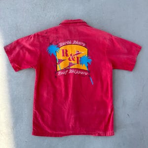Puede incluir: Camisa roja de manga corta con botones y un gráfico de palmeras y un pez marlin. El gráfico dice "North Shore R&J Reef Skippers".