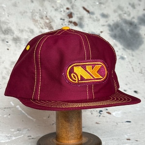 Pode incluir: Um boné de beisebol bordô com costuras amarelas em contraste e um botão amarelo na coroa. A parte frontal apresenta um emblema bordado com um logótipo estilizado em amarelo e bordô contendo as letras NK. A aba tem várias filas de costuras amarelas.