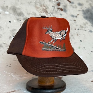 May include: A baseball cap featuring a dark brown brim and side panels with a bright orange front panel. The front panel displays a graphic illustration of a deer jumping over a fallen log in a forest setting, rendered in shades of brown and white.