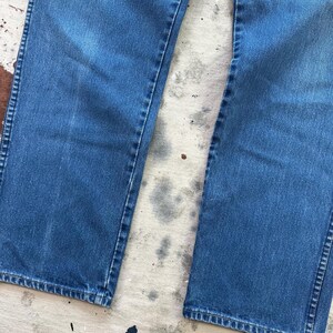 May include: Close-up of blue denim jeans. The jeans have a classic, straight-leg fit with visible seams and a faded appearance. The denim is a medium wash, showing signs of wear.