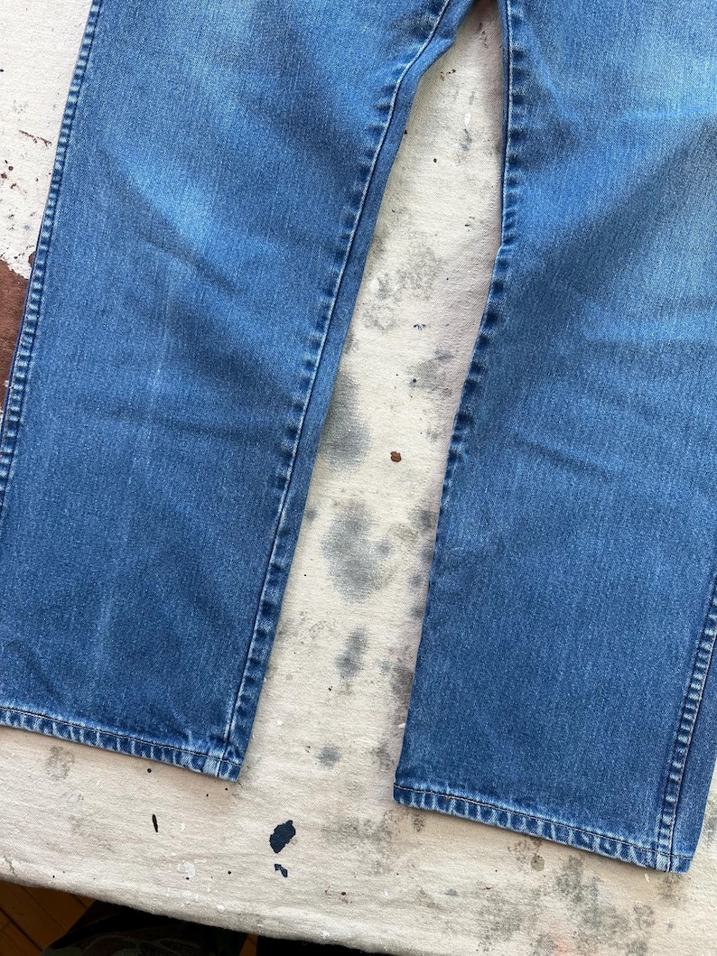 May include: Close-up of blue denim jeans. The jeans have a classic, straight-leg fit with visible seams and a faded appearance. The denim is a medium wash, showing signs of wear.