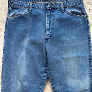 May include: Close-up of a pair of faded blue denim jeans. The jeans have a classic five-pocket design with a button and zipper closure. The denim shows signs of wear, with subtle fading and whiskering.