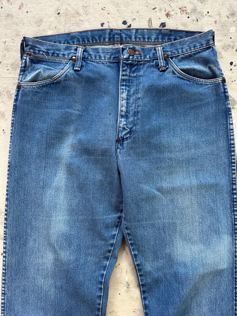 May include: Close-up of a pair of faded blue denim jeans. The jeans have a classic five-pocket design with a button and zipper closure. The denim shows signs of wear, with subtle fading and whiskering.