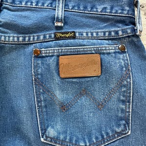 May include: Close-up of the back pocket of blue denim Wrangler jeans. The pocket has a classic design with a brown leather patch and the Wrangler logo. The jeans have a faded look, with visible stitching and metal rivets.