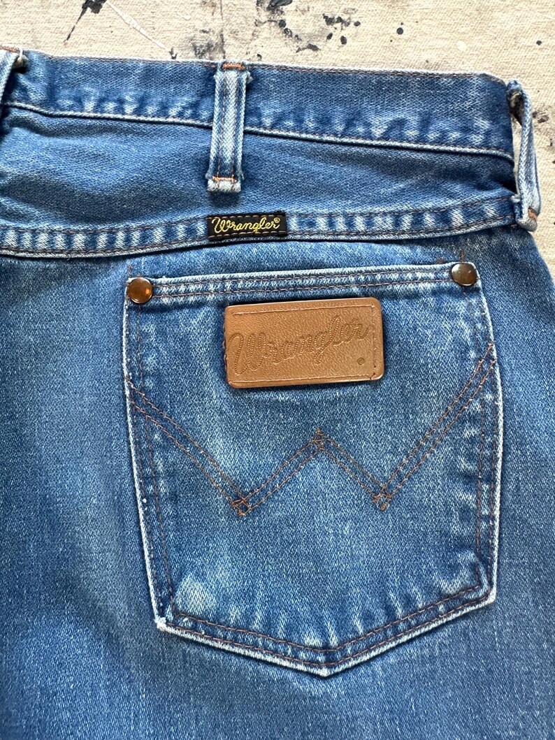 May include: Close-up of the back pocket of blue denim Wrangler jeans. The pocket has a classic design with a brown leather patch and the Wrangler logo. The jeans have a faded look, with visible stitching and metal rivets.