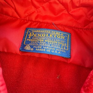 Puede incluir: Una prenda de color rojo brillante con una etiqueta de tela rectangular azul con texto dorado que dice: WARRANTED TO BE A PENDLETON, PENDLETON WOOLEN MILLS, PORTLAND, OREGON, 100% VIRGIN WOOL, L, PURE VIRGIN WOOL, MADE IN U.S.A. La tela es acolchada.