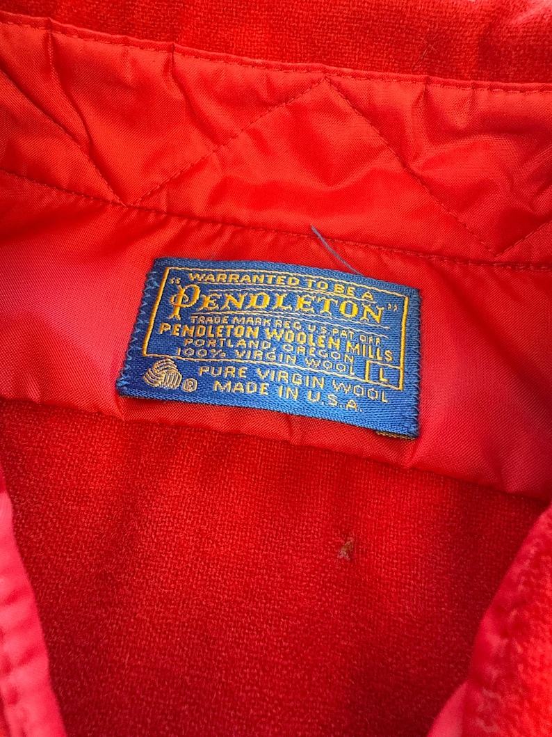 Puede incluir: Una prenda de color rojo brillante con una etiqueta de tela rectangular azul con texto dorado que dice: WARRANTED TO BE A PENDLETON, PENDLETON WOOLEN MILLS, PORTLAND, OREGON, 100% VIRGIN WOOL, L, PURE VIRGIN WOOL, MADE IN U.S.A. La tela es acolchada.