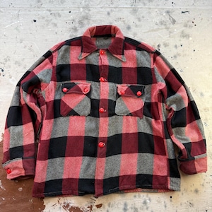 May include: A vintage, button-down shirt in a red, black, and grey plaid pattern. The shirt features two chest pockets with red buttons and a matching collar and cuffs. The shirt is made of a thick, woven fabric.