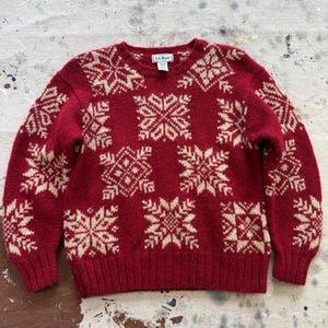 May include: A red crew neck sweater with a snowflake pattern in cream. The sweater has long sleeves and a ribbed hem and cuffs. The snowflake design is repeated across the front and sleeves. The L.L. Bean label is visible at the back of the neck.